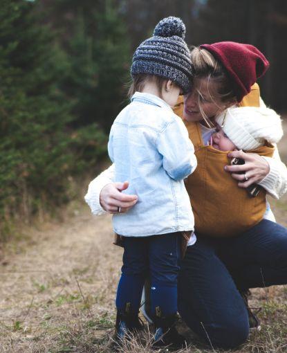 Mom with infant strapped to her chest kneels in front of a toddler.